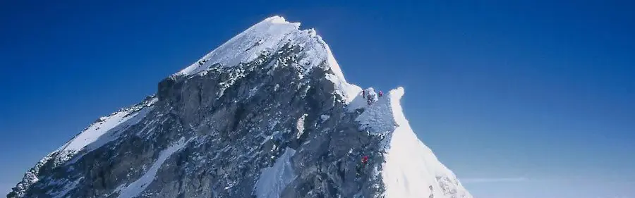 Een bergbeklimmer in een geel jack en klimuitrusting houdt een Nederlandse vlag vast op de besneeuwde top van een berg, met achter hem op de grond een rugzak en touwen.